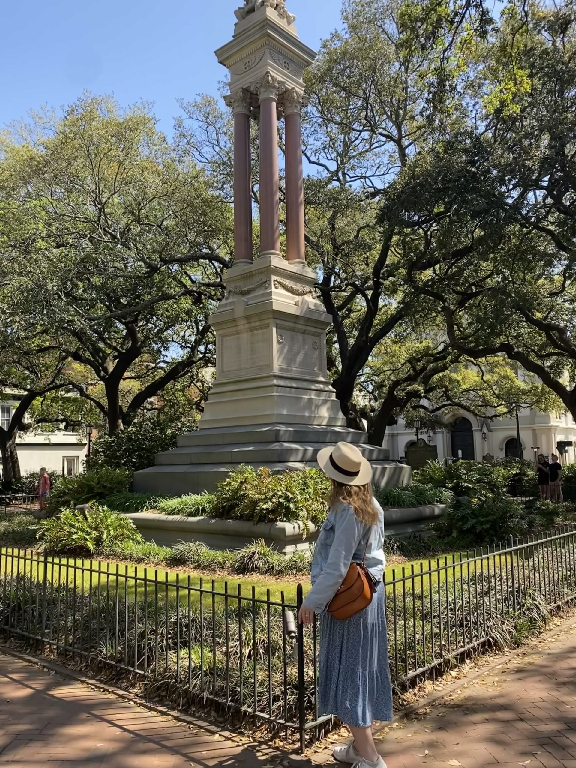 this is a photo of a woman wearing a hat and blue dress while standing in front of a monument in a square in Savannah, Georgia.